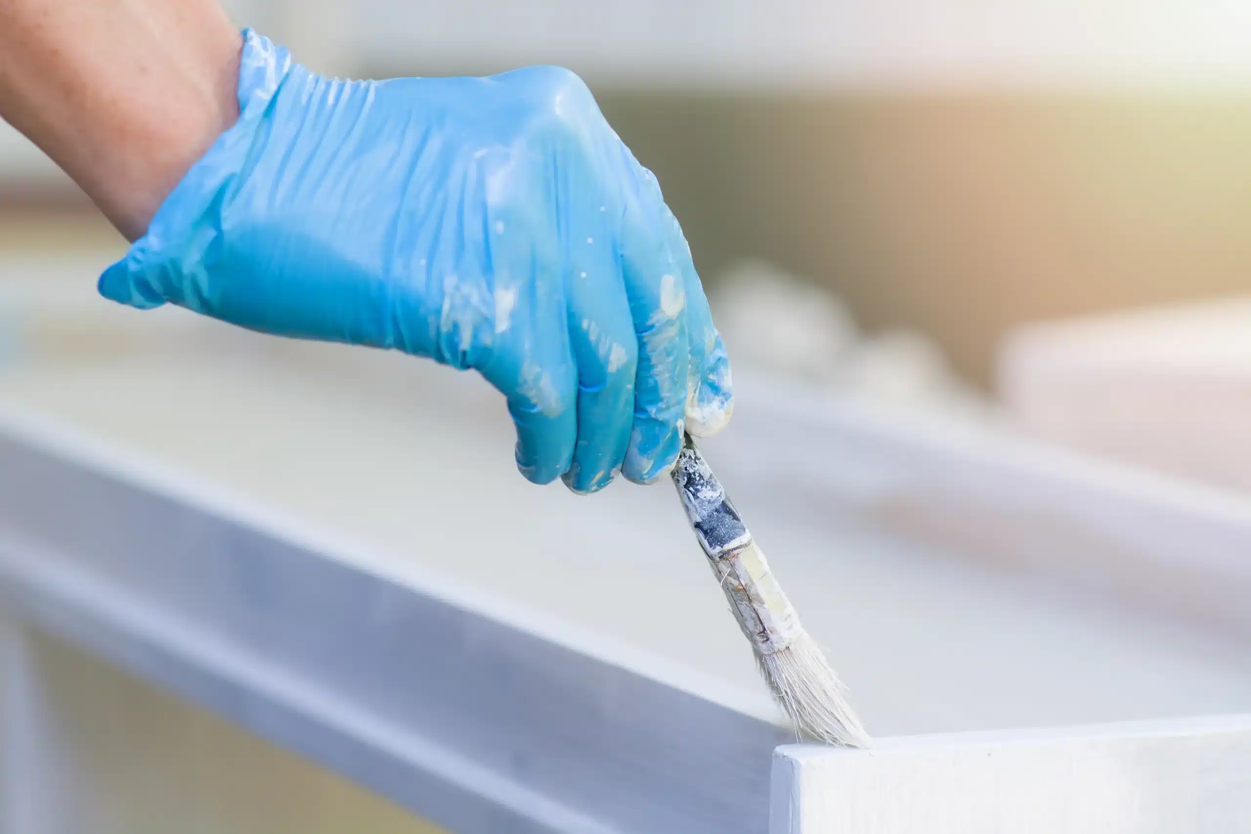 Hands in blue gloves hold a paintbrush and paint a new wooden plank with white paint outdoors. The man makes repairs. Close-up. The concept of construction, renovation, self-repair. What to be Prepared for When Painting Your Cabinets