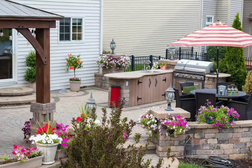 Summer flowers around an upscale brick patio with wooden gazebo, outdoor kitchen with BBQ and comfortable wicker dining furniture. right outdoor countertop